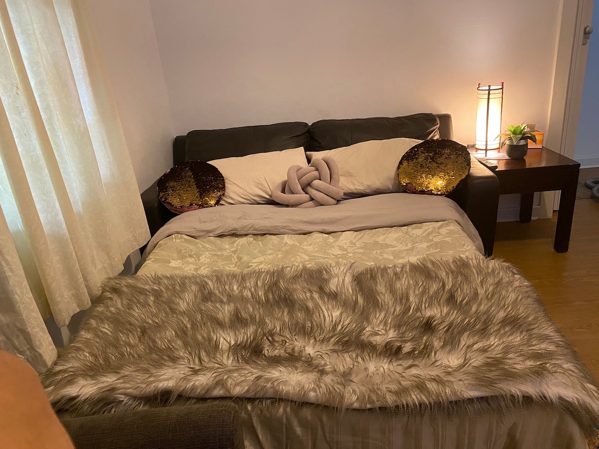 A comfortable bed is positioned against the wall, adorned with soft bedding and an elegant throw. Two decorative pillows complement the bed's aesthetic. A small table with a lamp is nearby, providing warm lighting, while sheer curtains allow gentle natural light to brighten the room.