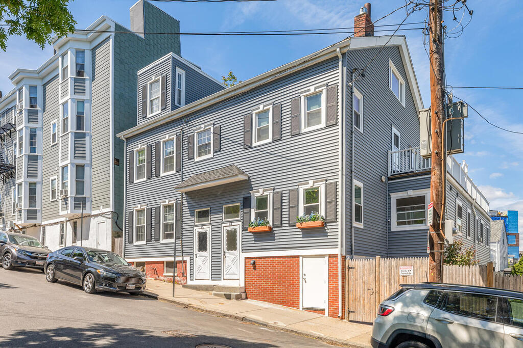The exterior of a two-story apartment building is visible, featuring gray siding and white trim. Planters with greenery are positioned by the windows. A porch with steps leads to the entrance, while a paved street runs along the front, showcasing parked vehicles.
