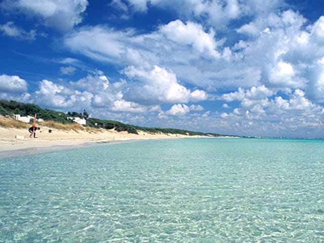 A sandy beach stretches along a calm, clear sea, with gentle waves lapping at the shore. The water is a gradient of light blue to turquoise, reflecting the bright sky dotted with fluffy white clouds. Greenery is visible in the distant background.
