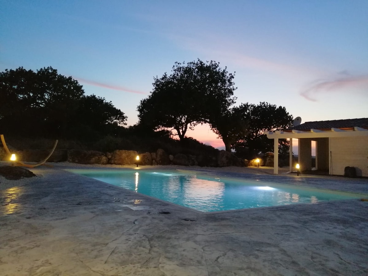 The image shows a tranquil outdoor area featuring a large swimming pool, illuminated gently by warm lights. Surrounding the pool are natural rock formations and trees, enhancing the serene atmosphere. A hint of sunset colors reflects on the water's surface, creating a calm setting.
