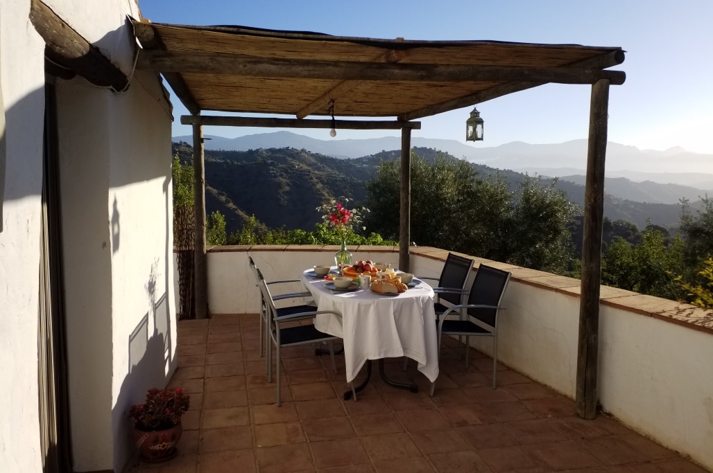An outdoor terrace features a round table set with a white tablecloth and an array of food items, surrounded by six metal chairs. A lantern hangs above, and panoramic mountain views are visible in the background, highlighting the natural beauty of the location.
