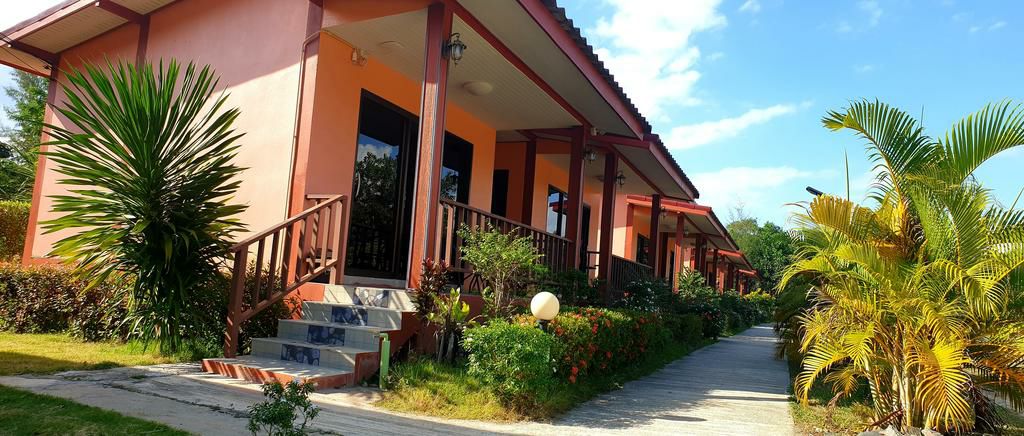 A pathway lined with vibrant greenery leads to a row of bungalows. Each bungalow features a covered porch with wooden railings and large windows, allowing natural light to illuminate the space. Surrounding plants enhance the inviting environment.