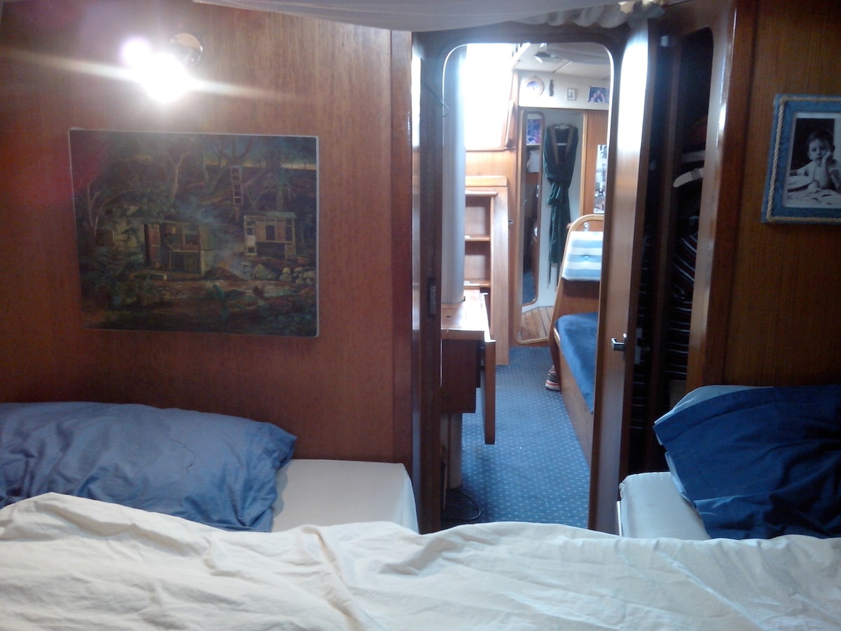 The image depicts a cozy interior of a boat, featuring a sleeping area with two beds adorned with blue pillows. A wooden paneling enhances the warm ambiance. A door leads to a bright living space with a table and seating area visible in the background.