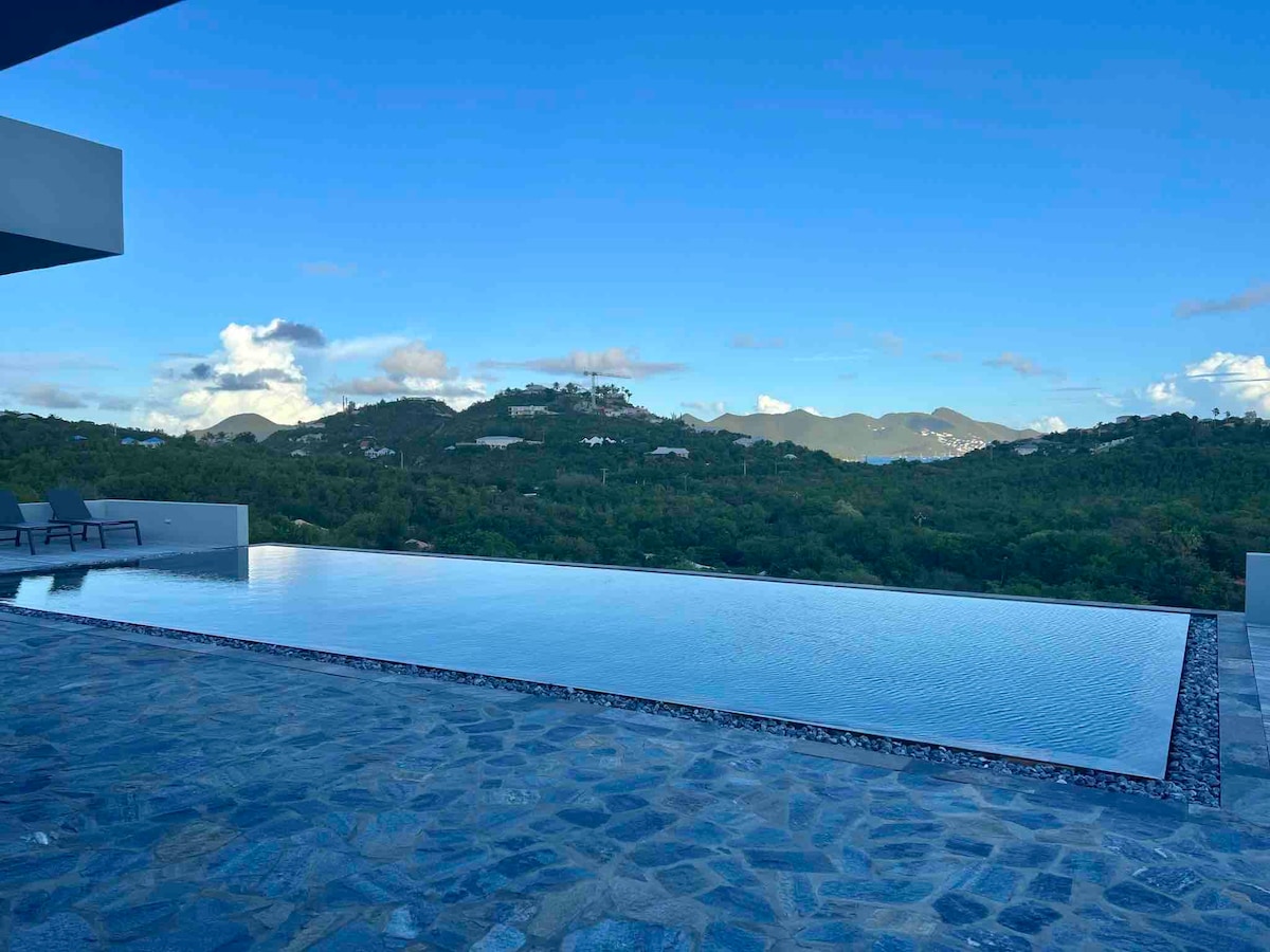 An infinity pool extends towards the horizon, merging with the lush greenery of the landscape. The tranquil water reflects the sky, while surrounding stone flooring complements the natural setting. Gentle hills are visible in the background, enhancing the peaceful ambiance.