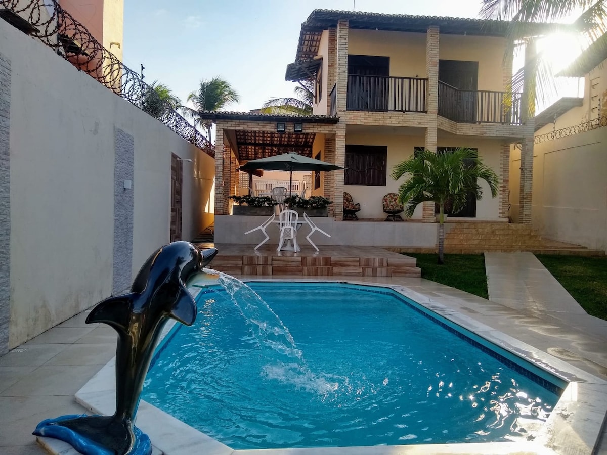 A serene pool area features a dolphin fountain, with clear water inviting relaxation. In the background, a two-story house showcases a blend of open spaces and shaded areas under a canopy. Lush greenery surrounds the poolside, enhancing the tranquil atmosphere.