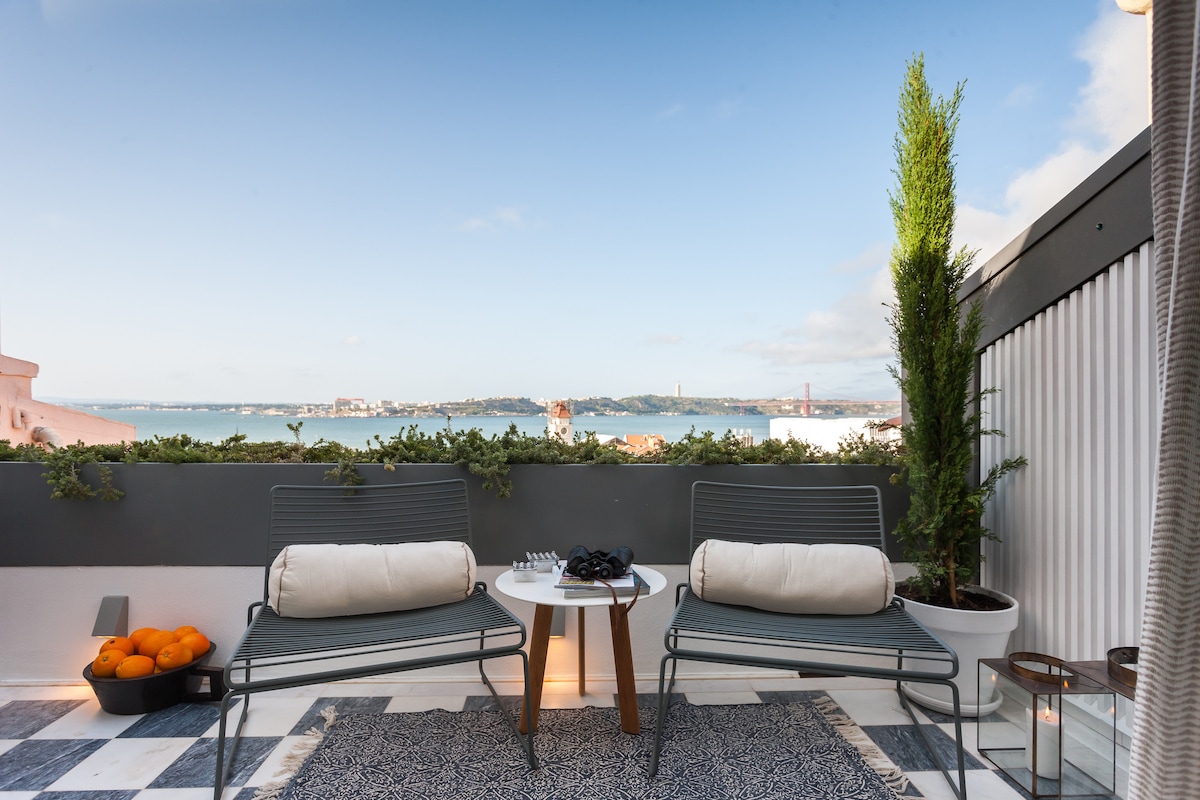 A private terrace features two modern chairs positioned beside a small table. The area is surrounded by greenery, offering sweeping views of the Tagus River and Lisbon's skyline. A bowl of oranges is nearby, and soft lighting enhances the relaxing ambiance.