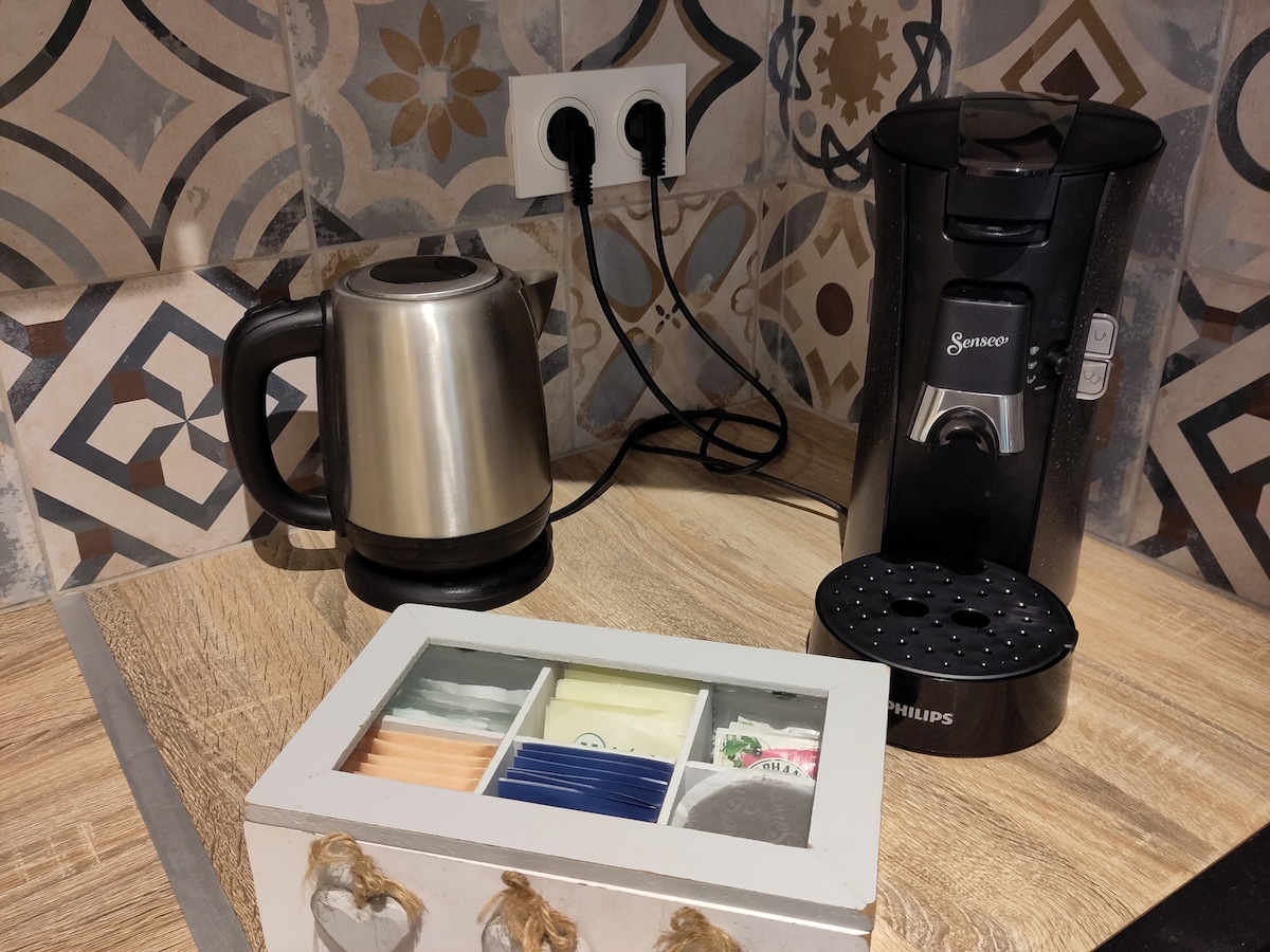 A kitchenette area is presented, featuring a silver kettle and a black coffee machine. Nearby, a decorative box holds an assortment of tea and coffee packages, showcasing various colors and labels. Unique patterned tiles create an appealing backdrop.