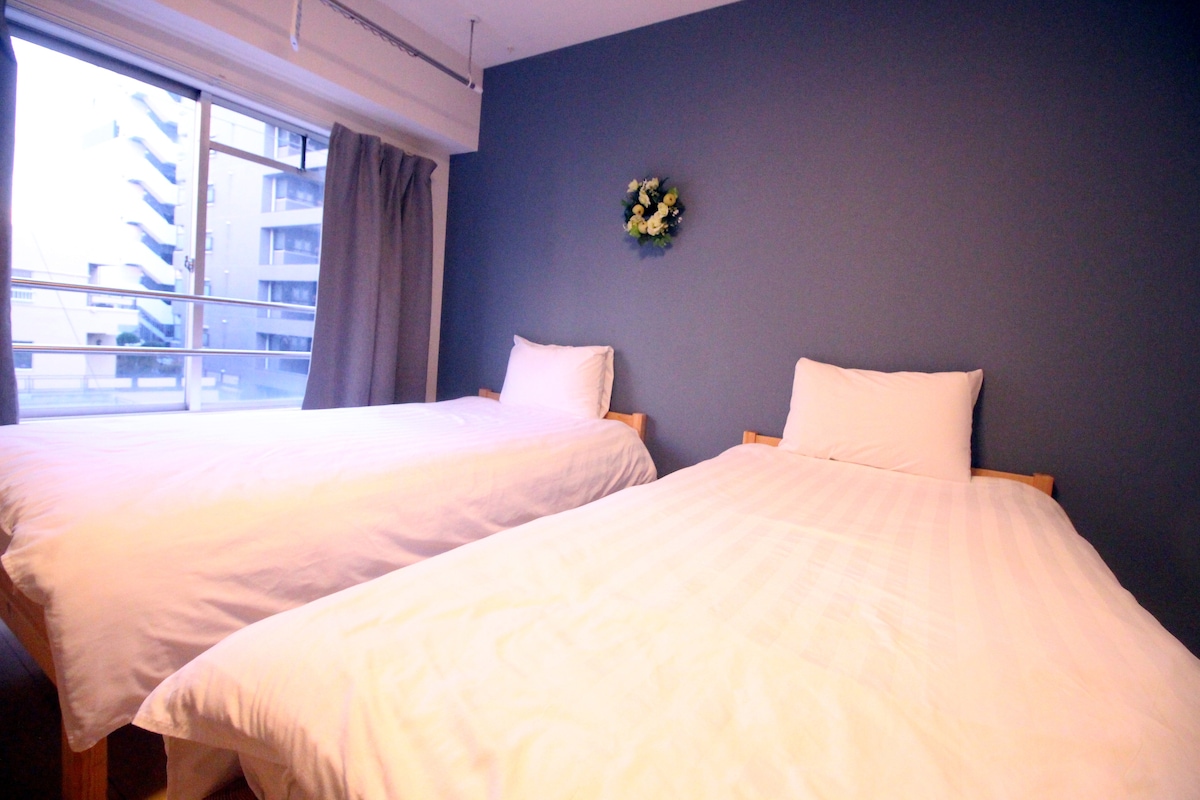 Two spacious beds are positioned against a blue wall, each adorned with white linens and decorative pillows. Natural light is allowed in through large glass doors, which lead to a balcony. A floral wall decoration adds a subtle touch.