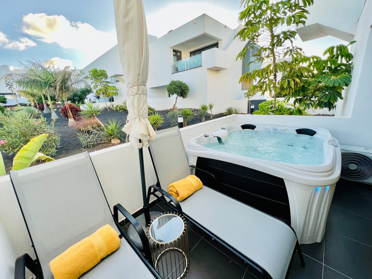 A private terrace features a jacuzzi surrounded by lush garden views. Two lounge chairs are complemented by soft yellow towels, while a small table provides a convenient surface. The space offers a serene setting for relaxation, with modern architecture visible in the background.