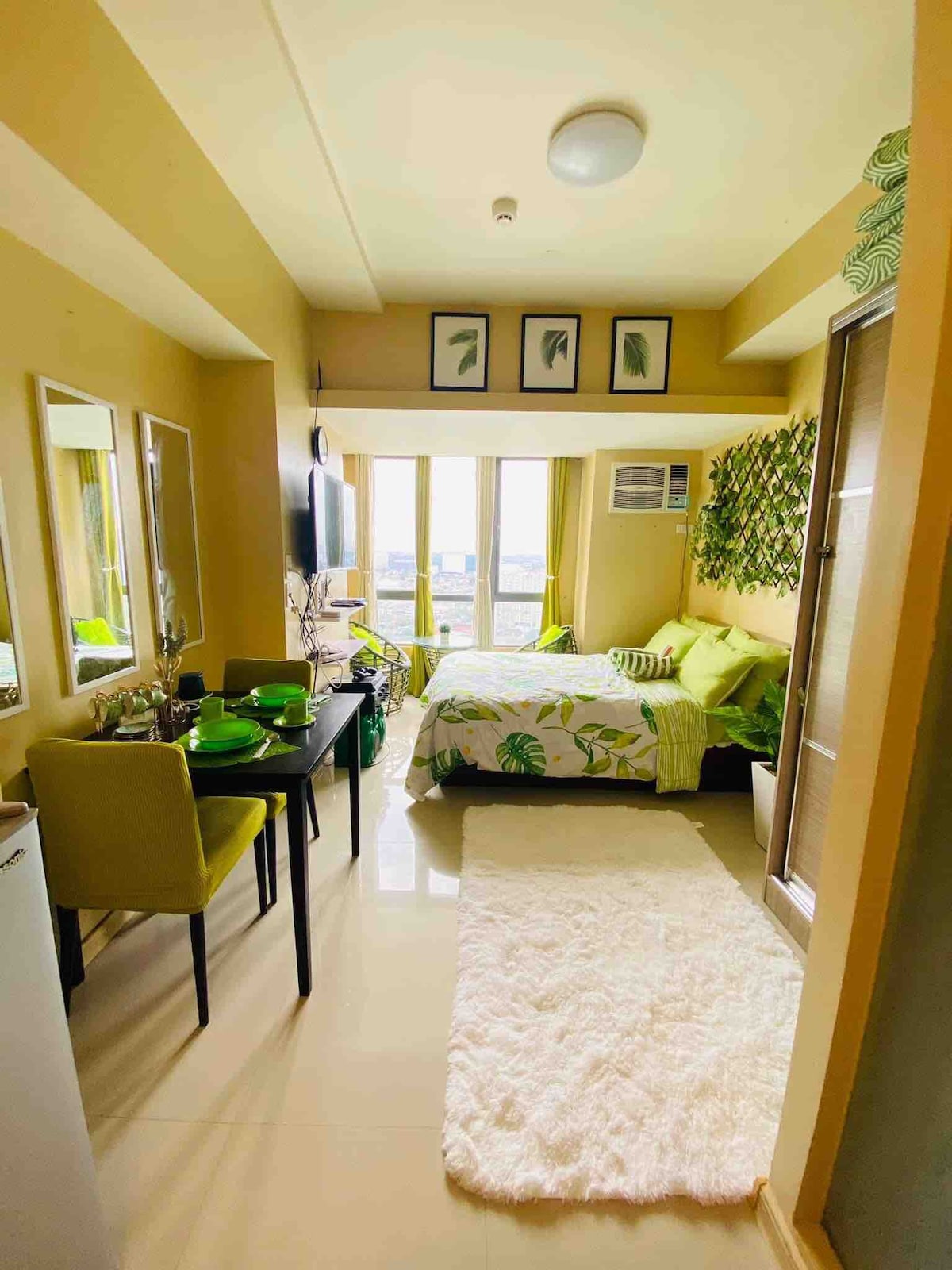 A vibrant interior showcases a cozy sleeping area with greenery-themed decor, complemented by a spacious bed covered in light-colored bedding. A small dining table with chairs is positioned near the window, which allows natural light to brighten the room. Full-length mirrors add depth to the space.