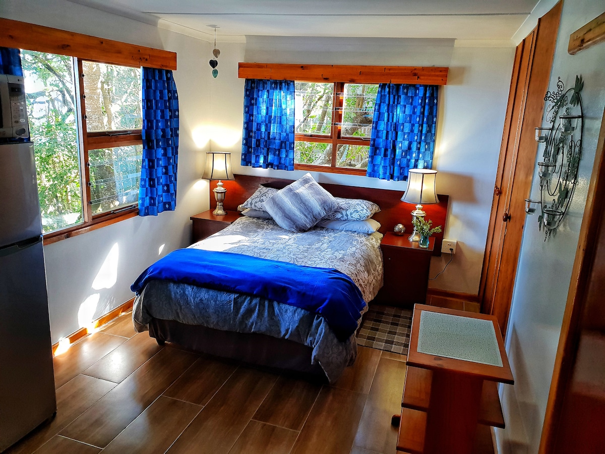 A cozy sleeping area features a spacious bed with blue accents and multiple pillows. Large windows provide natural light, framed by blue curtains. Nightstands with lamps are positioned on either side. A refrigerator is visible in the corner, adding convenience to the room.