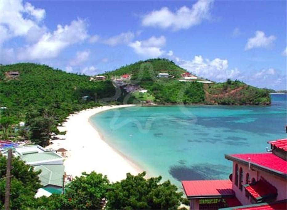 A panoramic view of a serene beach curves along the coastline, surrounded by lush green hills. The crystal-clear water reflects the blue sky, dotted with fluffy clouds. Beachfront properties and palm trees are visible, inviting relaxation in a tranquil setting.