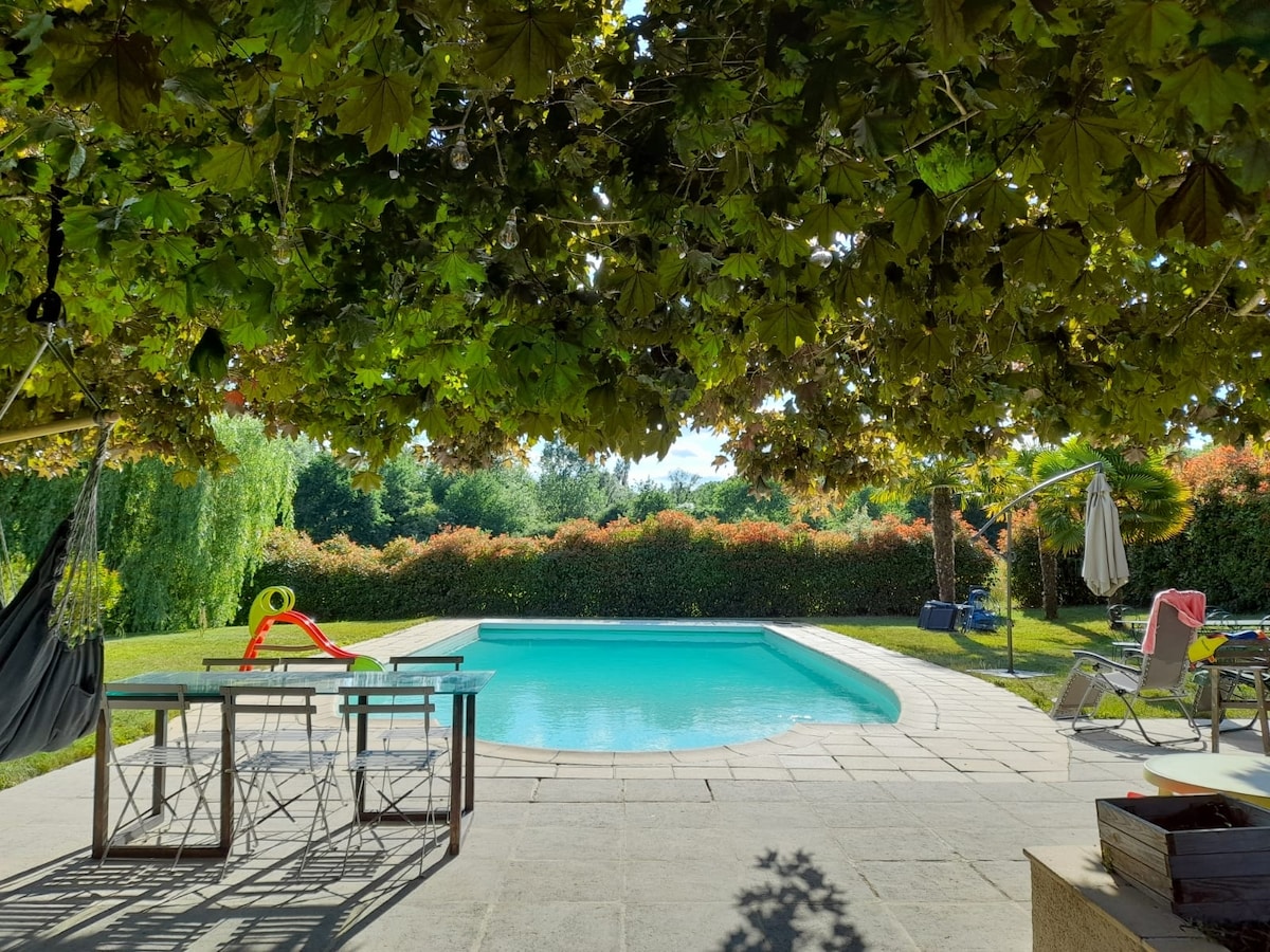 An inviting outdoor area features a swimming pool surrounded by lush greenery. A shaded dining table sits under a canopy of leaves, offering comfort for meals. Lounge chairs and a hammock provide additional relaxation options, contributing to a serene ambiance.
