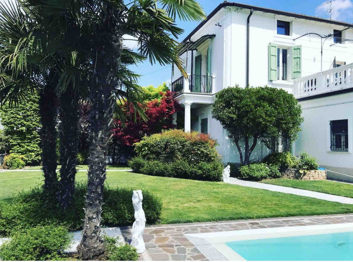 A spacious garden surrounds a villa, featuring well-maintained greenery and vibrant flower beds. Palm trees are positioned nearby, and two white sculptures are visible on the lawn. The villa, with its white exterior and green shutters, reflects a serene and inviting environment.