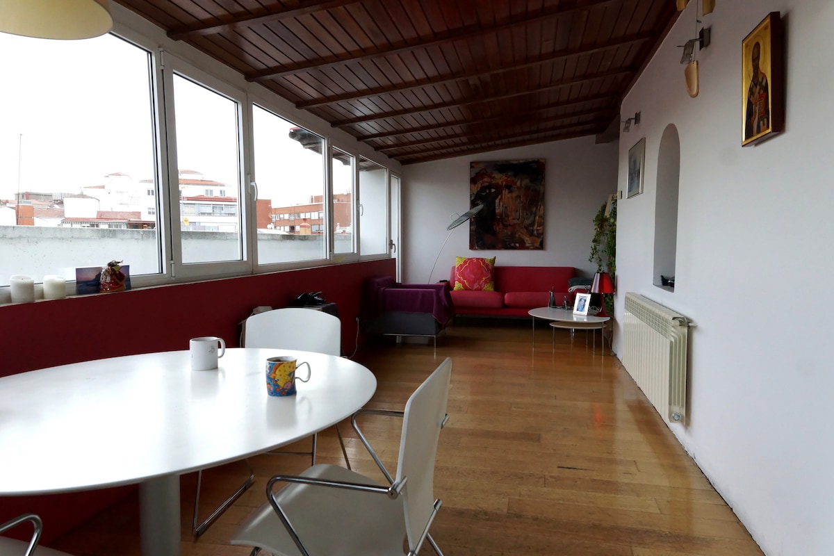 A spacious living area features a wooden ceiling and multiple large windows allowing for natural light. A red sofa is complemented by a purple throw, and a round dining table with four chairs occupies the space. Decorative artwork and a plant add character to the room.