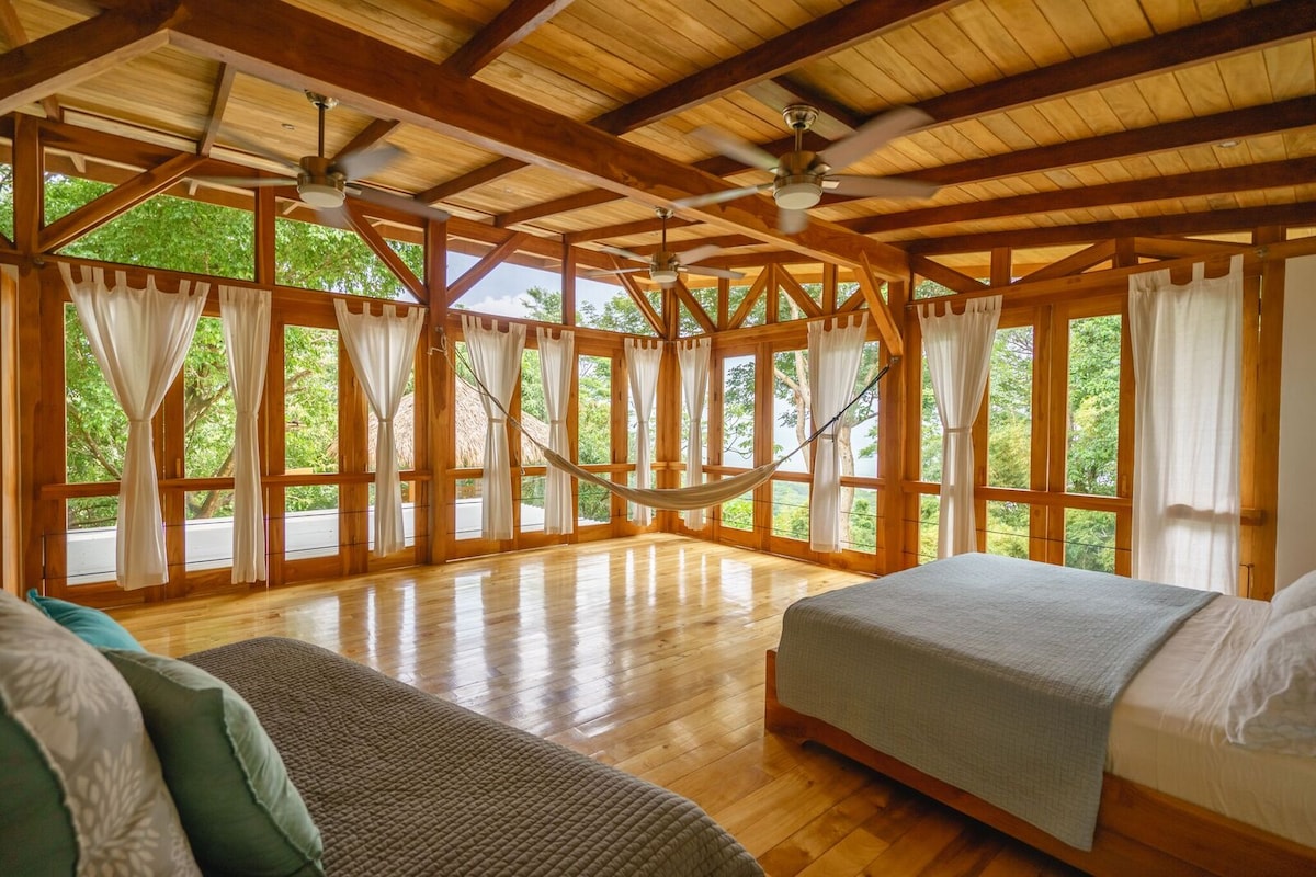 A spacious bedroom is highlighted by wooden beams and large windows that provide views of the surrounding greenery. Two beds, adorned with light-colored bedding, are positioned on polished wood flooring. Soft curtains frame the windows, and a hammock is visible, enhancing the serene atmosphere.
