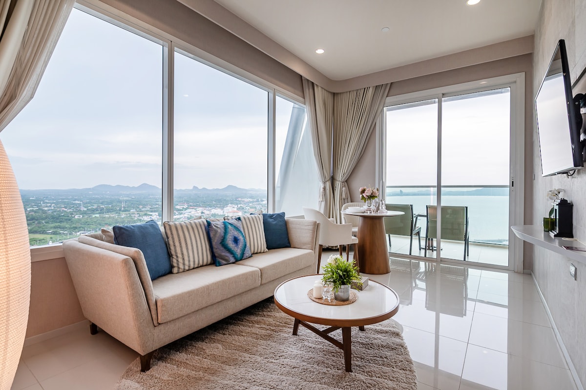 A modern living area features large windows offering expansive views of the coastline. A light-colored sofa with decorative cushions is positioned alongside a wooden coffee table. A small dining table with chairs is situated near the balcony, enhancing the space's open, airy feel.