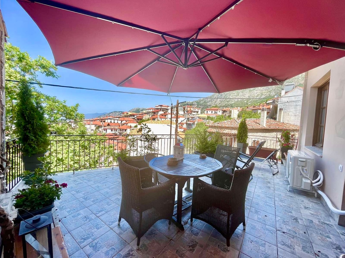 A spacious terrace is set with a round dining table surrounded by wicker chairs. A large umbrella provides shade, while flowers in planters add a touch of greenery. Scenic views of the village and surrounding mountains can be seen in the background.