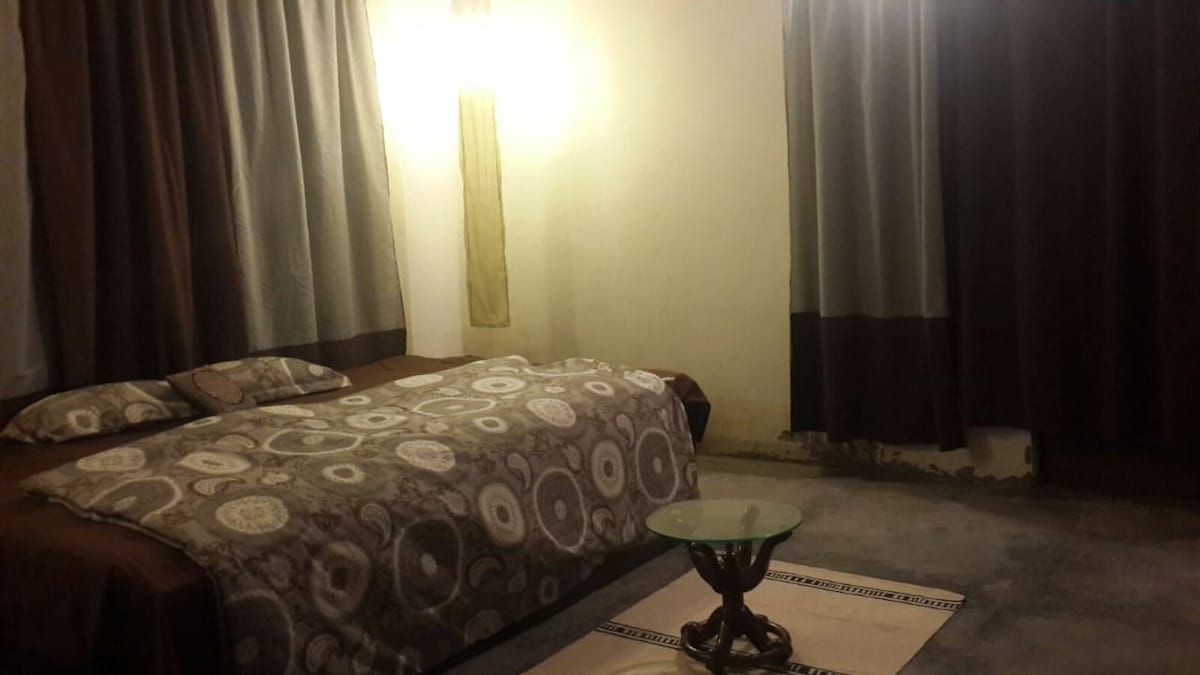 A spacious sleeping area is featured, with a large bed covered in a patterned blanket and bolstered by soft pillows. Light fixtures gently illuminate the room, complemented by floor-to-ceiling curtains in neutral tones. A decorative table sits on the floor, enhancing the overall setting.