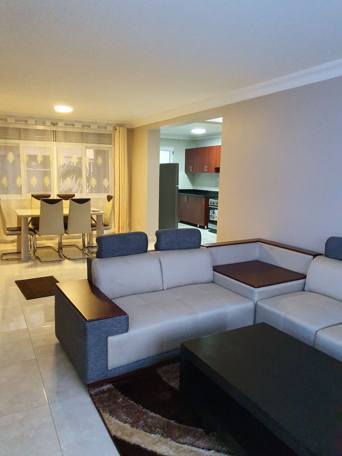 A spacious living area showcases a modern sectional sofa with wooden accents, positioned next to a dark coffee table. A dining area with light-colored chairs is visible in the background, leading to a well-equipped kitchen. Soft lighting and neutral-colored walls create a comfortable atmosphere.