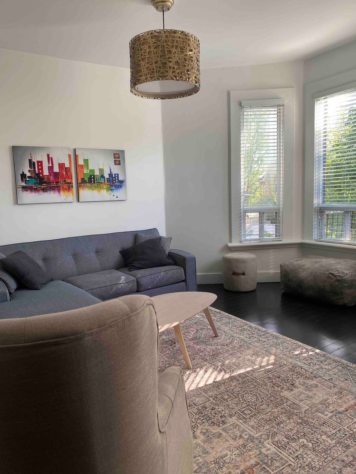A well-furnished living area features a gray sofa complemented by two soft cushions. A wooden coffee table sits in front, with an area rug below. Large windows allow natural light to fill the space, and a piece of colorful art adorns the wall.