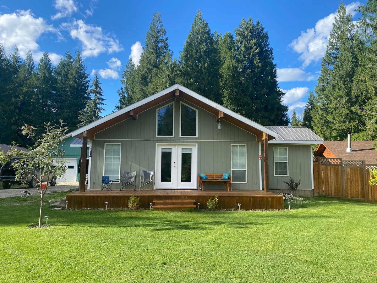 The exterior of a modern cabin features a sloped roof and large front windows. A wooden deck extends from the entrance, accompanied by a seating area. Lush green grass surrounds the cabin, complemented by tall trees in the background under a clear blue sky.