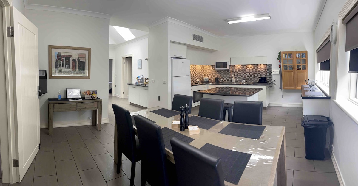A spacious kitchen and dining area features a large dining table set for six with dark chairs. Modern appliances are integrated into the sleek cabinetry, and a mosaic tile backsplash adds a subtle touch of texture. Ample natural light enters through windows, enhancing the open layout.