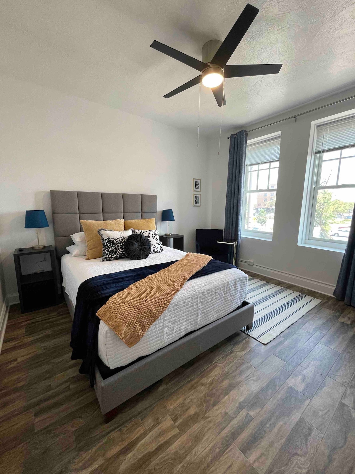 A queen bed with a padded headboard is positioned against a bright wall. Complementary pillows are arranged neatly on the bed. Natural light enters through two large windows adorned with dark curtains. A ceiling fan and two bedside lamps are visible, enhancing the cozy ambiance.