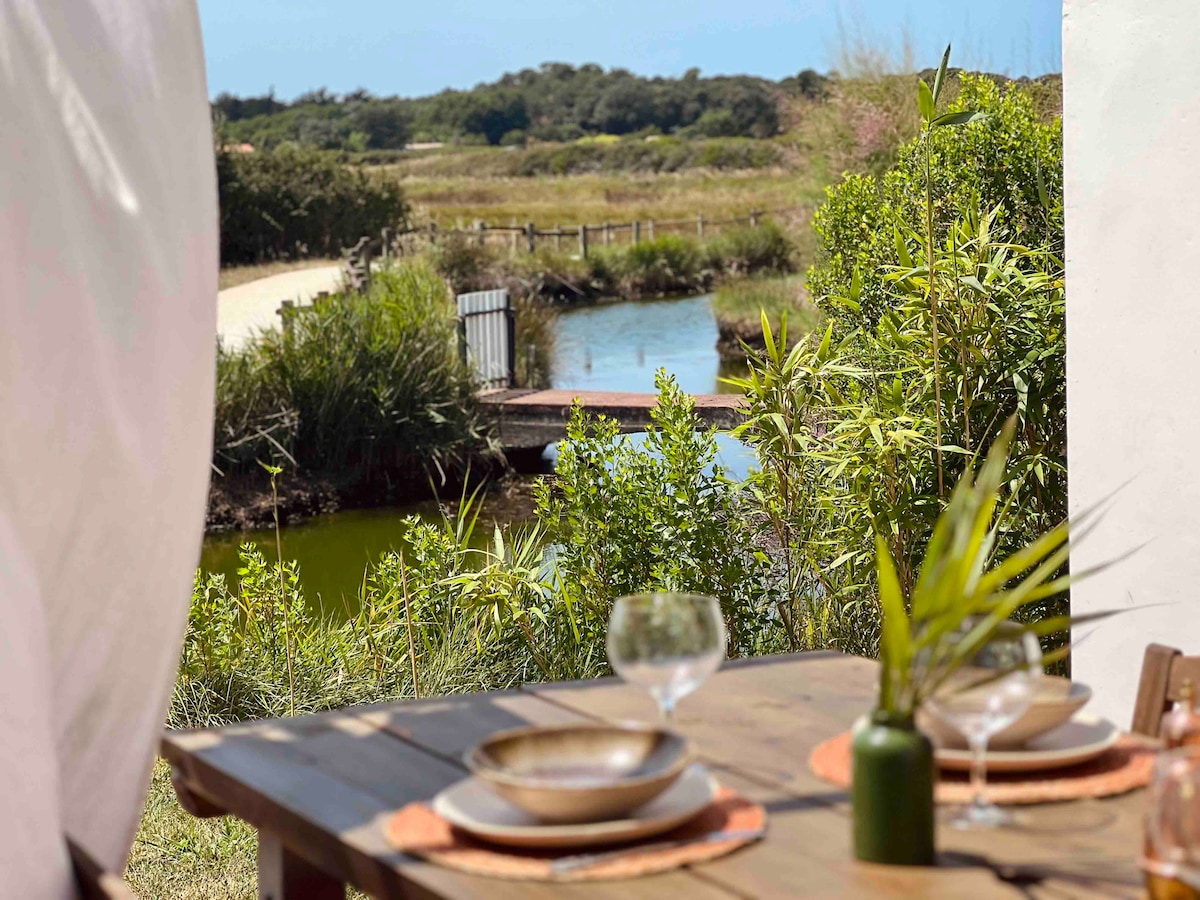 A wooden dining table is set for two, featuring plates and glasses. A serene view of lush greenery and a tranquil waterway can be seen in the background, with a footbridge crossing the waterway, surrounded by natural landscapes.