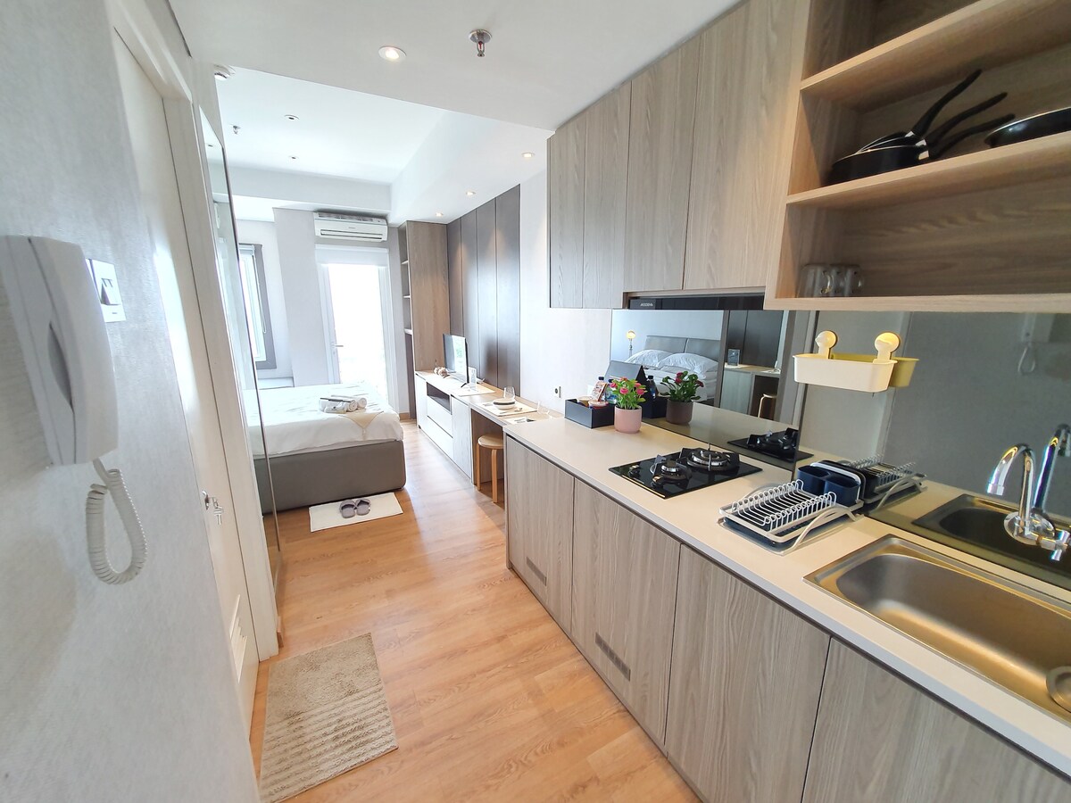 The open layout features a compact kitchen with modern cabinetry, equipped with cooking utensils and appliances. A cozy sleeping area is visible in the background, highlighted by natural light from the window. Wood laminate flooring runs throughout, adding warmth to the space.