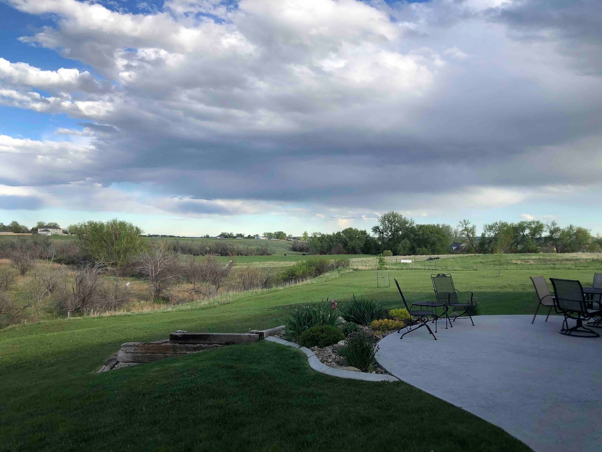 A spacious patio area is viewed from above, featuring a circular concrete surface surrounded by outdoor seating. Lush green grass leads to a view of open fields and scattered trees under a partly cloudy sky, creating a serene outdoor atmosphere.