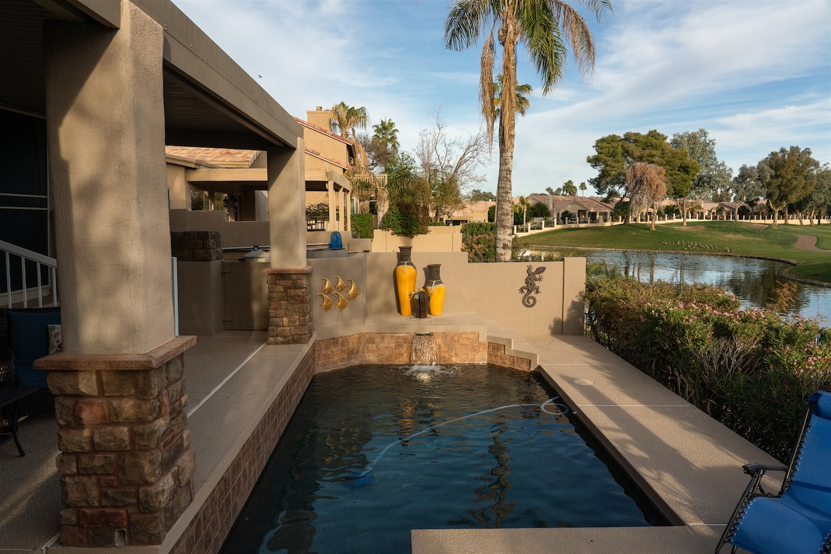 An outdoor patio area features a stone-accented spa spilling water into a small pool. Colorful decorative elements, including yellow pots, are arranged artfully around the space. A lush green golf course and trees are visible in the background under a clear sky.