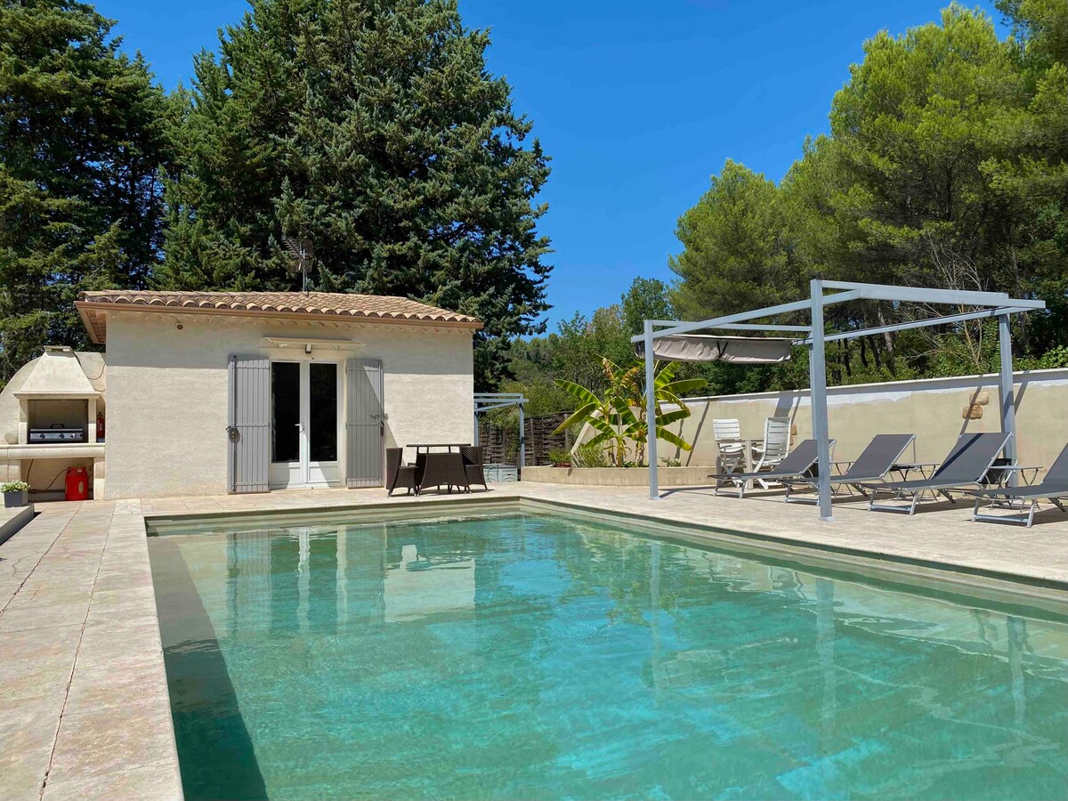 A clear view of the shared swimming pool surrounded by a sun deck is presented. Comfortable lounge chairs are positioned under shaded areas, and a barbecue setup is visible nearby. The pool area is complemented by a light-colored building with doors that open directly onto the patio.