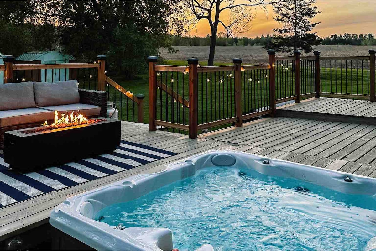 An outdoor deck features a luxurious hot tub and a fire table surrounded by a wooden railing. A cozy seating area is arranged with plush cushions. The background includes open fields and a sunset sky, creating a serene atmosphere.