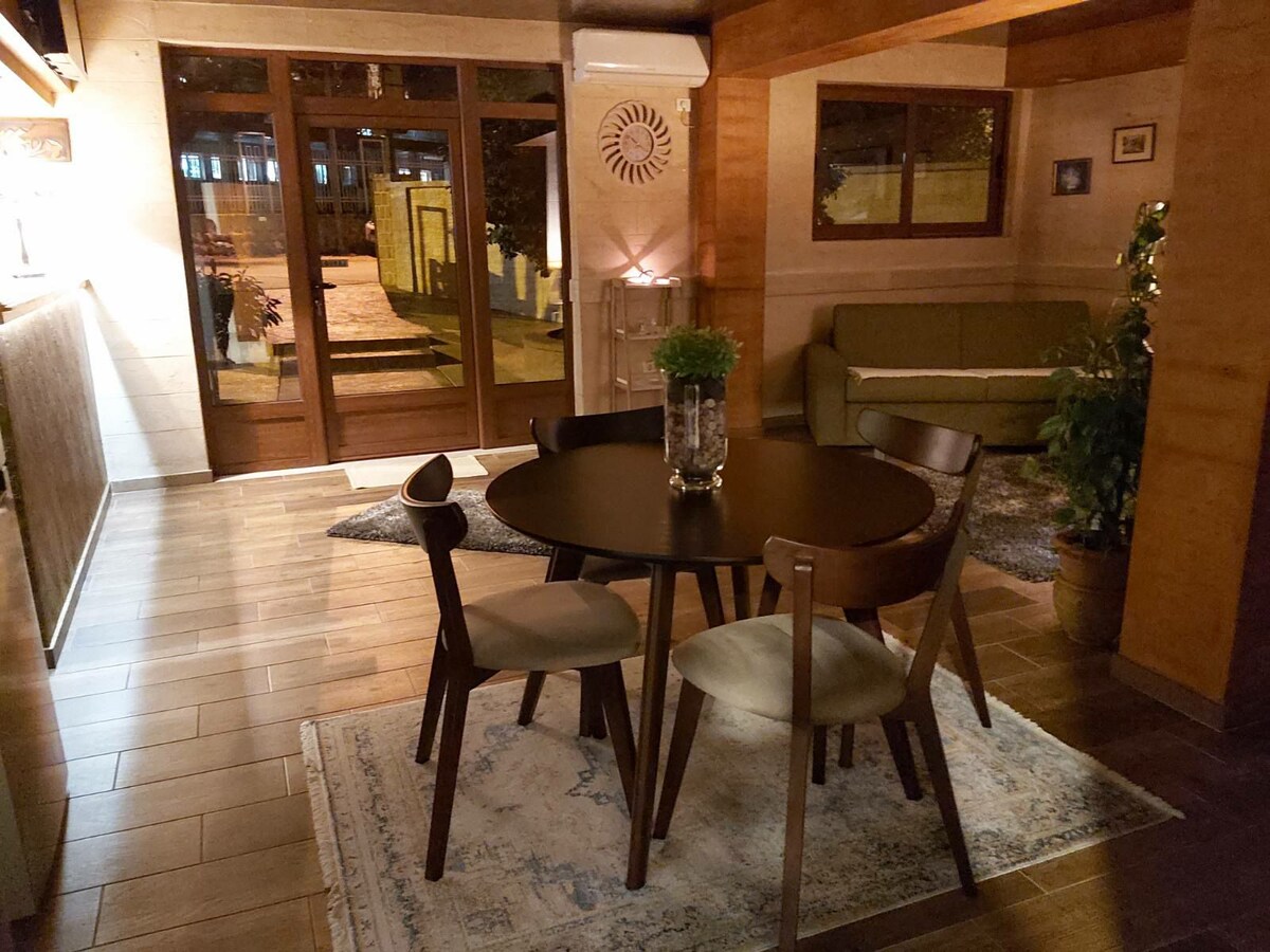 A modern dining area features a round table surrounded by four wooden chairs. A soft rug is placed beneath the table, and a small plant sits in a glass vase at its center. Large windows allow natural light, with a view of the outdoor space.