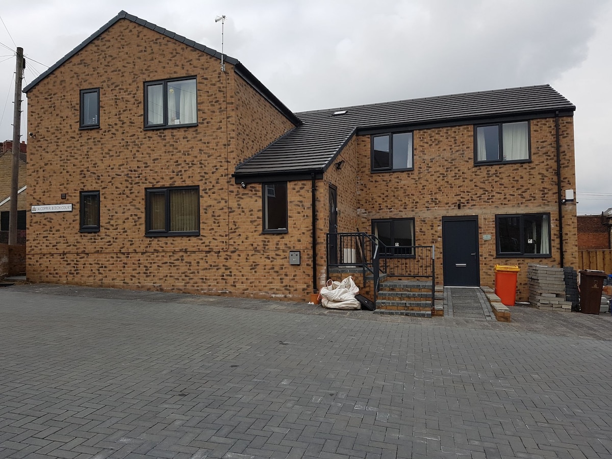 The exterior of a newly built apartment block is presented, featuring a combination of brick and large windows. The ground level includes a staircase leading to the entrance, while a paved area provides ample space for parking. The setting appears quiet and residential.