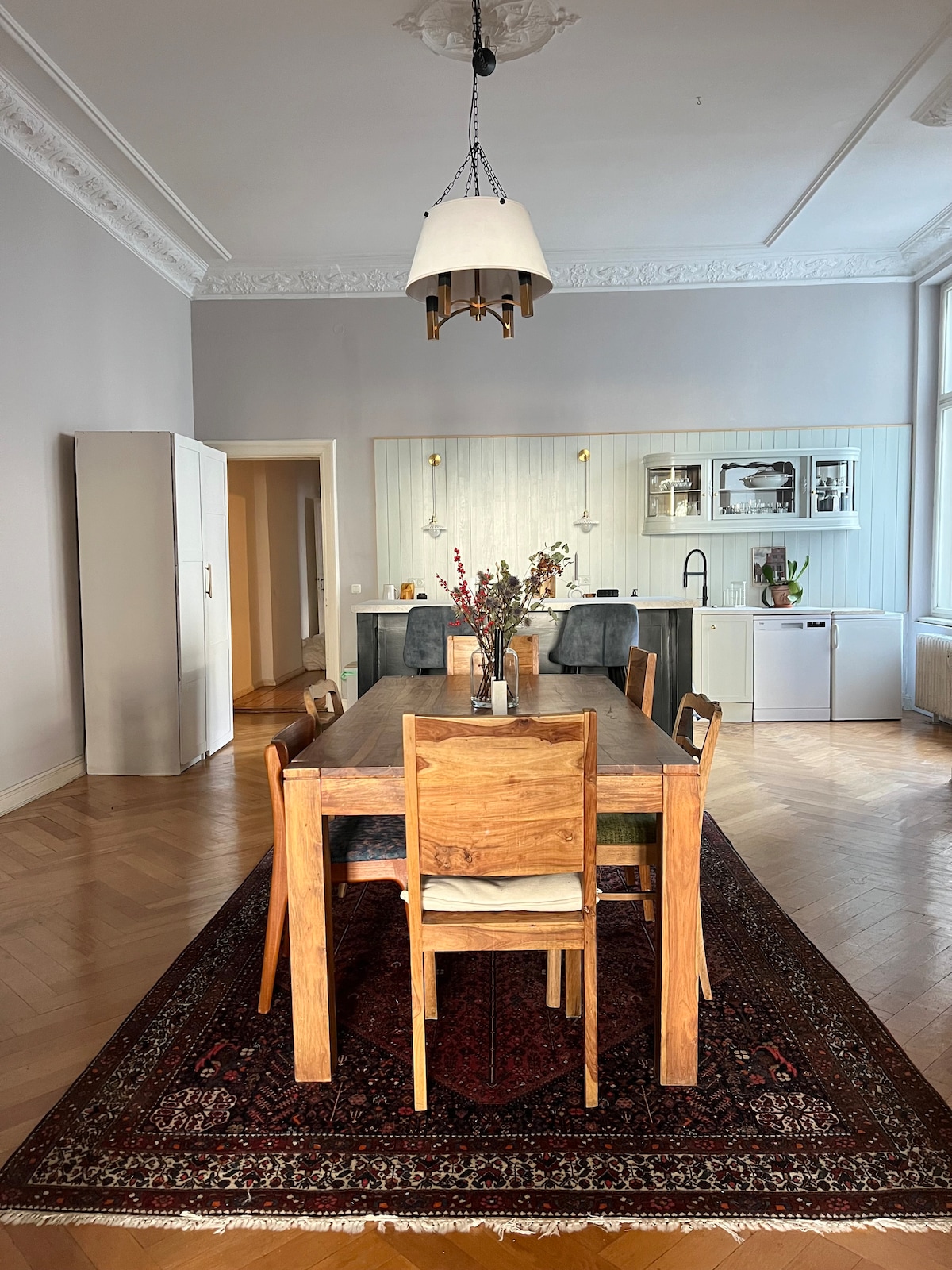 A spacious dining area is illuminated by a pendant light. A long wooden table is surrounded by six chairs, with a decorative runner adding warmth. A large mirror reflects the room, while an open kitchen is visible in the background, equipped with modern appliances.