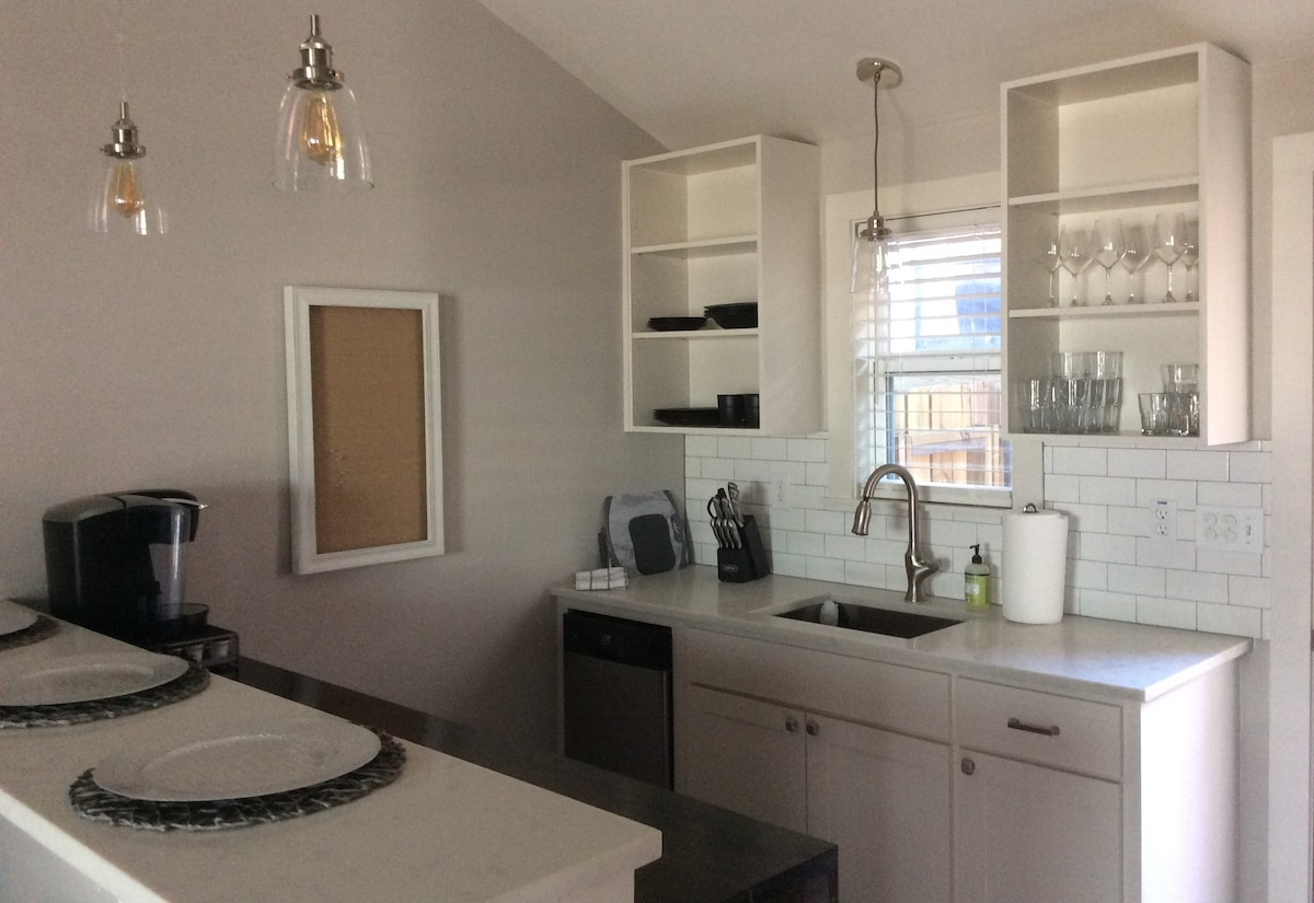 The kitchen features modern cabinetry with open shelving displaying glassware and dishware. A stainless steel sink is accompanied by a contemporary faucet, while a coffee maker is positioned next to the counter. Natural light enters through a window adorned with blinds.