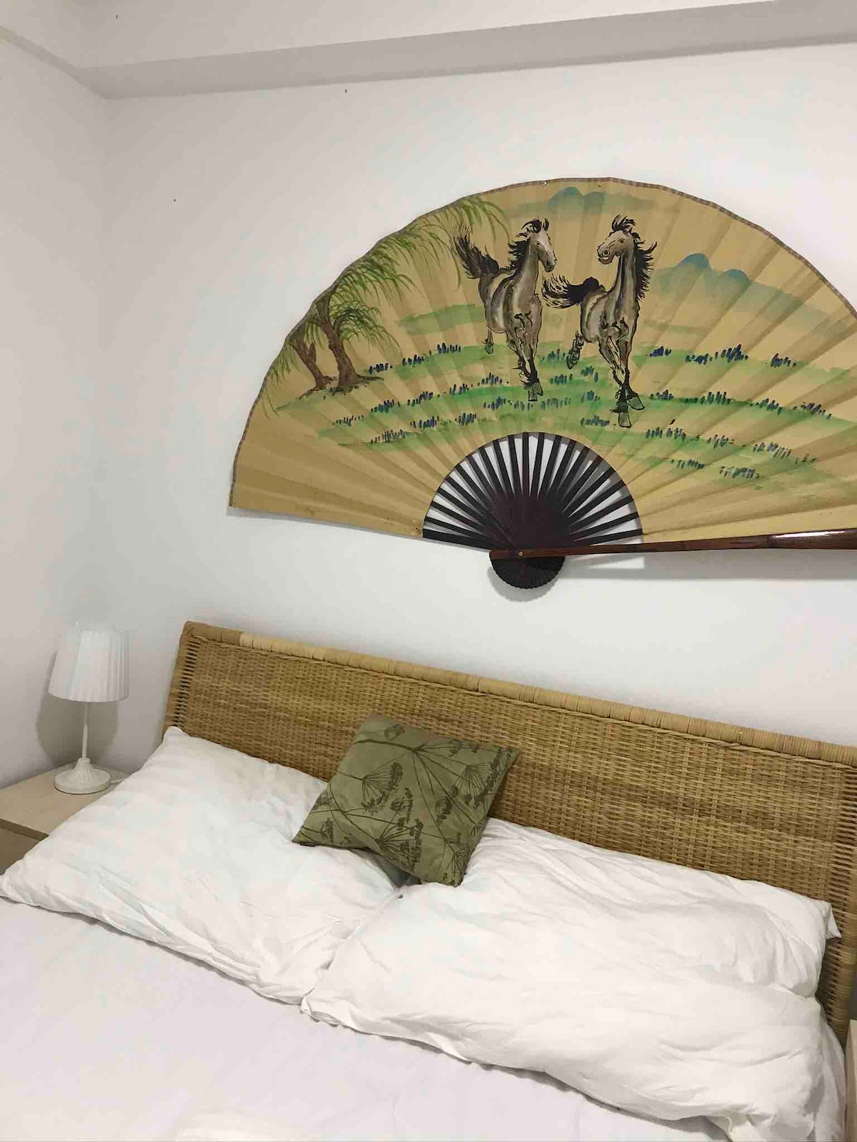 A spacious bed with crisp white linens is positioned against a neutral wall. Above the headboard, an ornate fan displays a serene landscape with two figures. A bedside lamp stands to the left, providing soft lighting.