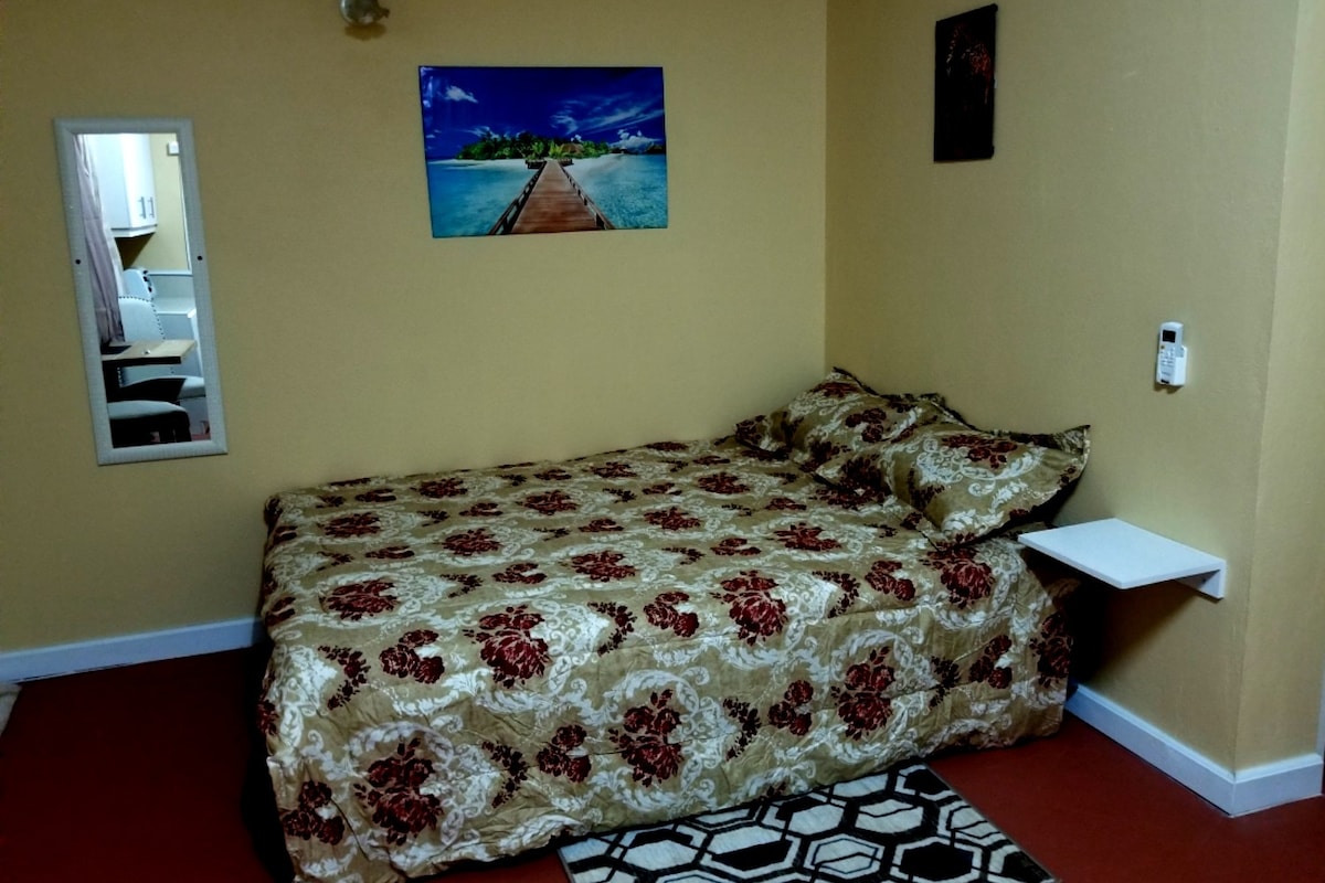 A studio apartment features a double bed with floral bedding against a yellow wall. A small wall-mounted table is visible beside the bed. A mirror and an artwork depicting a tropical landscape add to the decor. A patterned rug completes the cozy space.