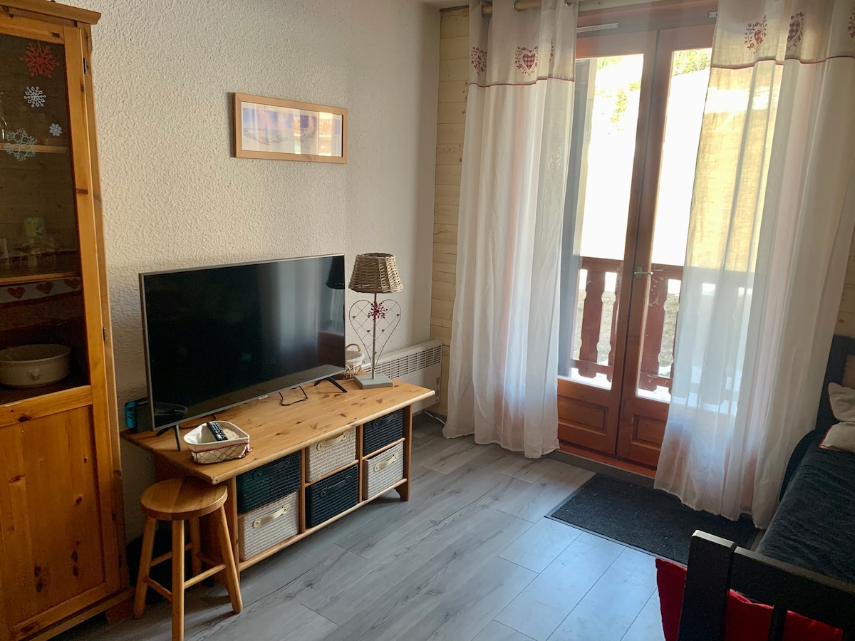 A living area features a wooden TV stand with a flat-screen television and a small stool. Light flows through double doors leading to a balcony, framed by sheer curtains. A decorative lamp and wall art add subtle touches to the space.