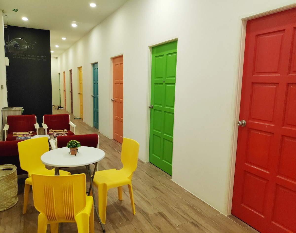 A brightly lit corridor features colorful doors in shades of pink, blue, green, yellow, and red. The space includes a small round table surrounded by yellow chairs, complemented by red and white seating arrangements nearby.