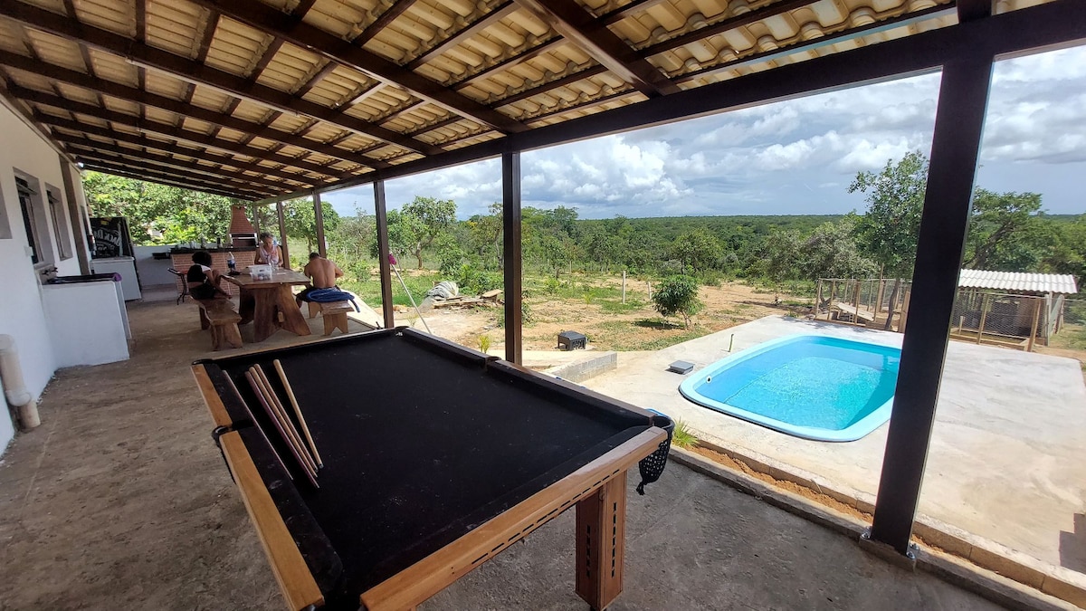 An outdoor patio area features a pool table, with a view of a spacious backyard and swimming pool in the distance. Natural vegetation surrounds the space, providing a serene backdrop. The covered structure allows for comfortable outdoor seating, inviting social activities amidst nature.