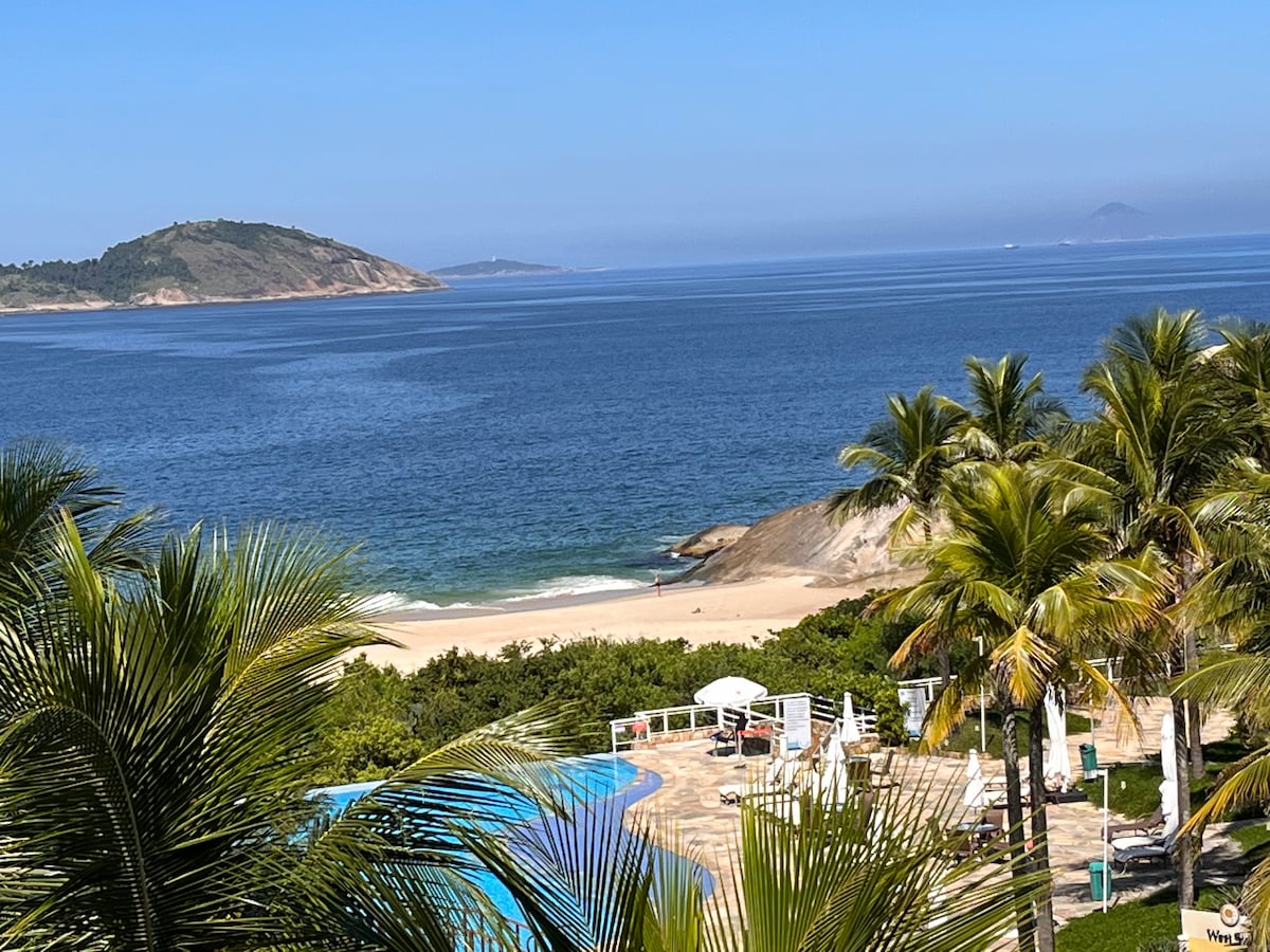 The image displays a serene coastal view featuring gentle waves lapping at a sandy beach. Lush palm trees frame the foreground, while an expansive blue sea stretches to the horizon, complemented by distant hills and a clear blue sky.