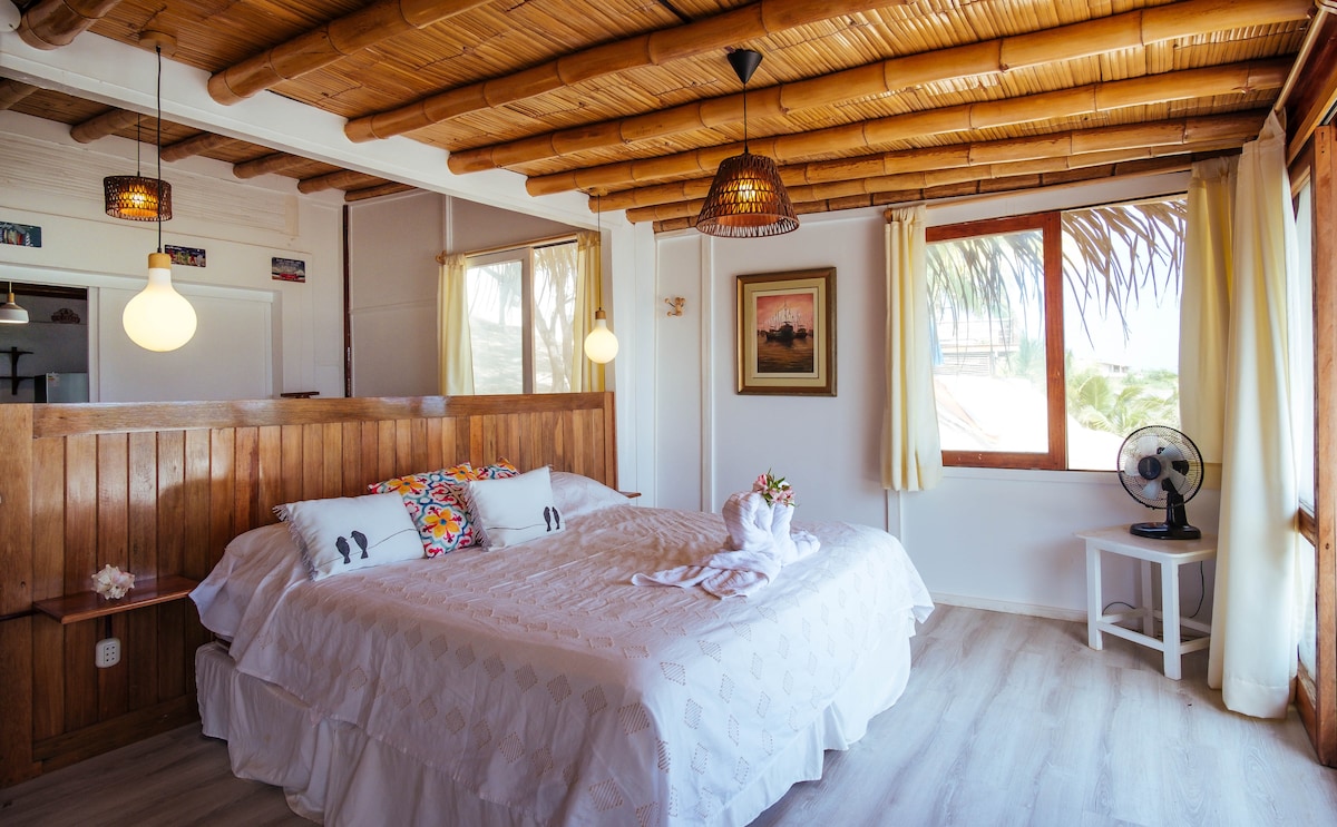 A cozy bedroom features a spacious bed adorned with white linens and decorative pillows. Natural light filters through a large window, complemented by light curtains. A ceiling with bamboo details and warm pendant lights create a serene atmosphere, while a fan provides additional comfort.