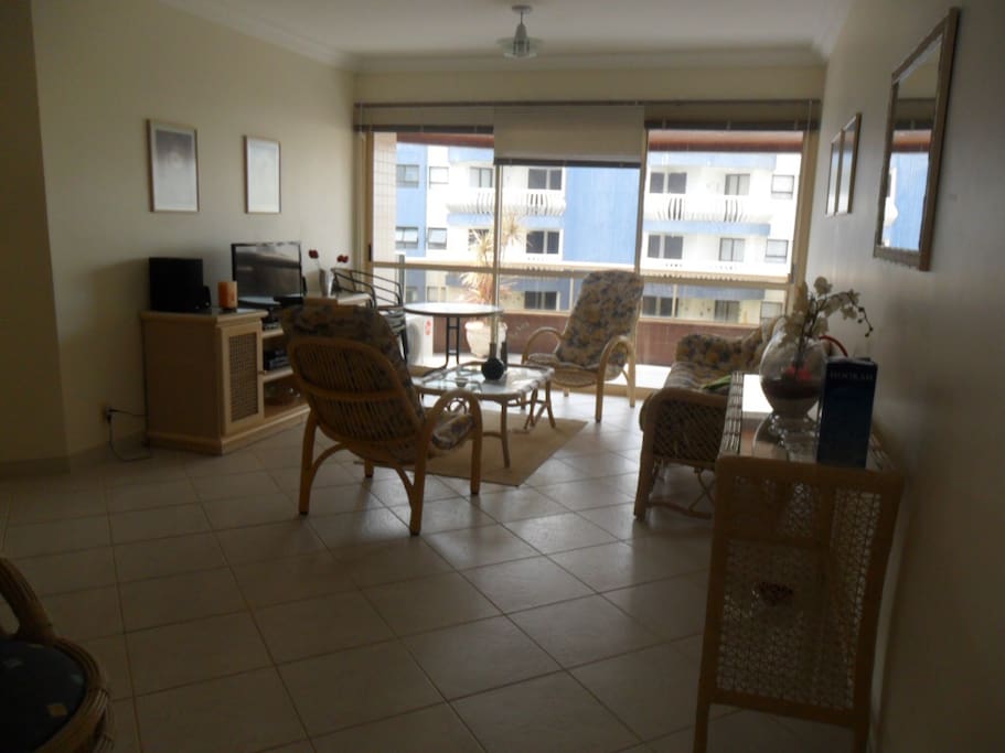 Vista da entrada do apartamento. Ampla sala de estar com mesa de jantar para 4 pessoas, vista para a varanda e uma mesa com 4 cadeiras para a varanda