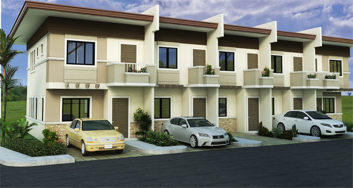 A row of contemporary townhouses features a clean, modern design with decorative stone accents. Each unit includes a small balcony and windows for natural light. Vehicles are parked in front, indicating easy access to each door, while green landscaping adds a welcoming touch to the exterior.