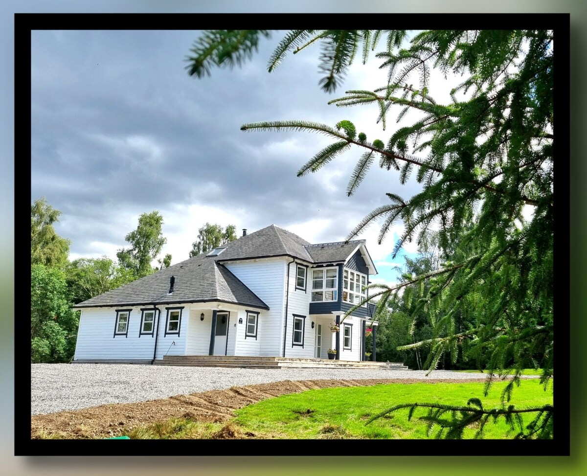 The exterior of a spacious white villa is captured, framed by greenery. The house features multiple gables and large windows, surrounded by a well-maintained gravel driveway and lush lawn. The cloudy sky adds a serene atmosphere to the setting.