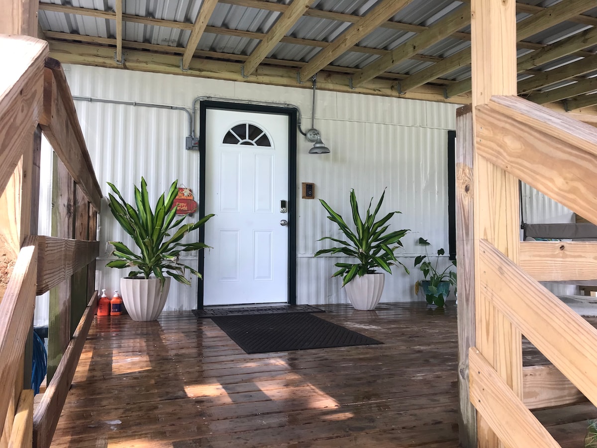 The entrance features a white door framed by two formal planters with green foliage. A welcome mat is positioned in front of the door, while the wooden deck is supported by sturdy beams overhead, providing a shaded area.