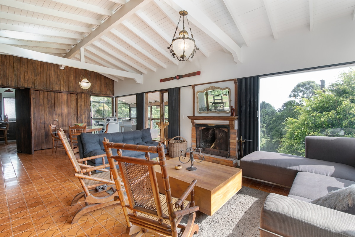 A spacious living area is highlighted by a large window that frames views of the surrounding greenery. A rustic wooden ceiling contrasts with the light walls. Comfortable seating includes a dark sofa, rocking chairs, and a coffee table centered around a fireplace, creating an inviting space.